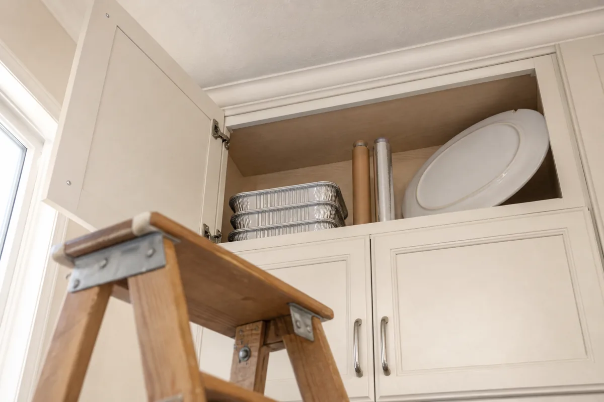 Dramatic low-angle view looking up at the top of a high upper cabinet with the kitchen ceiling visible above; the open top shelf holds only a short stack of disposable foil roasting pans, a roll of kraft parchment paper and a roll of aluminum foil standing upright side-by-side, and one large oval matte-white holiday serving platter leaned vertically against the back wall — a wooden two-step stepladder sits in the foreground on the kitchen floor, slightly out of focus, establishing that the only way to reach this shelf is to climb up