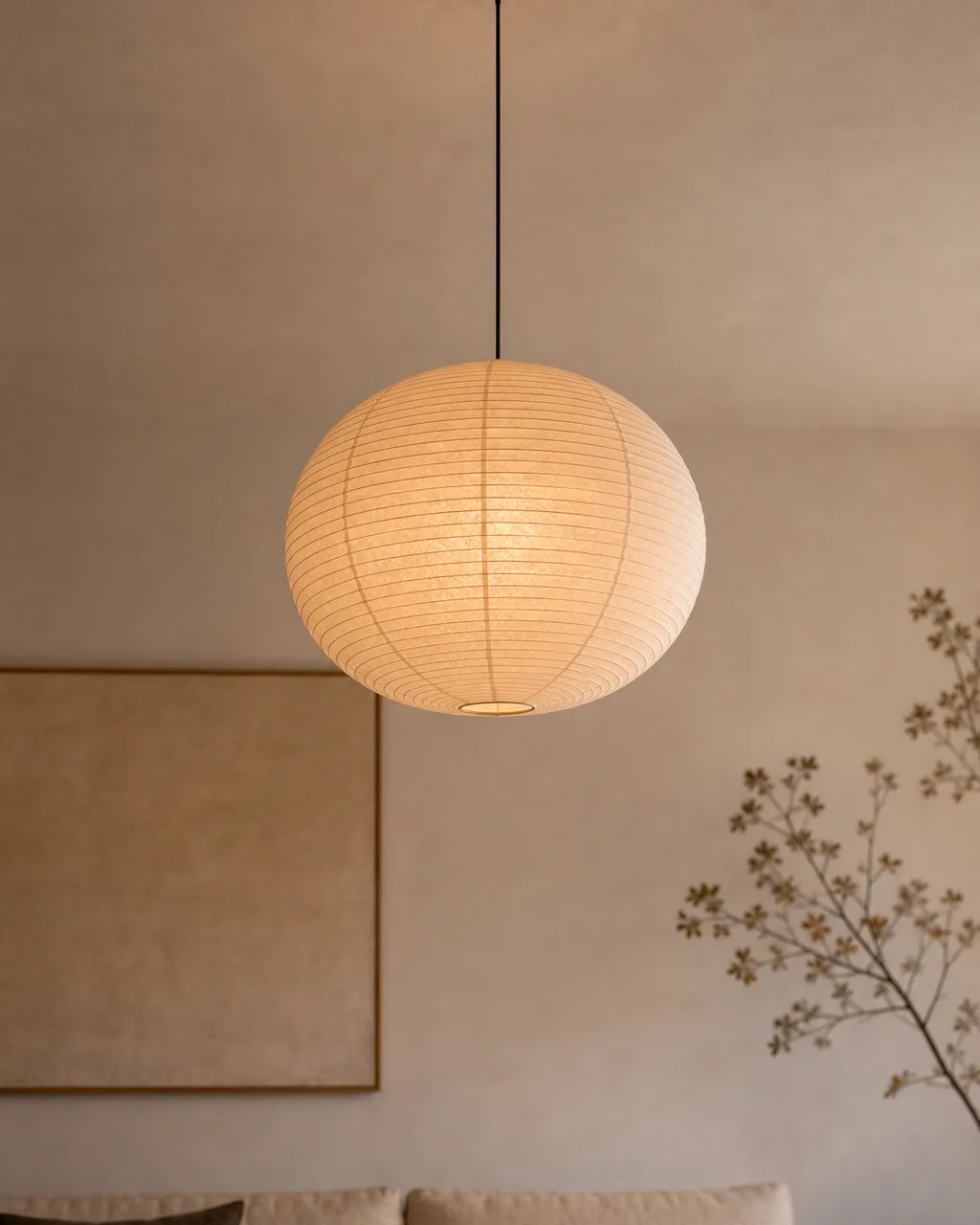 A large round washi-style paper pendant lamp in pale cream hanging from a white ceiling, casting a warm glow over a sofa below
