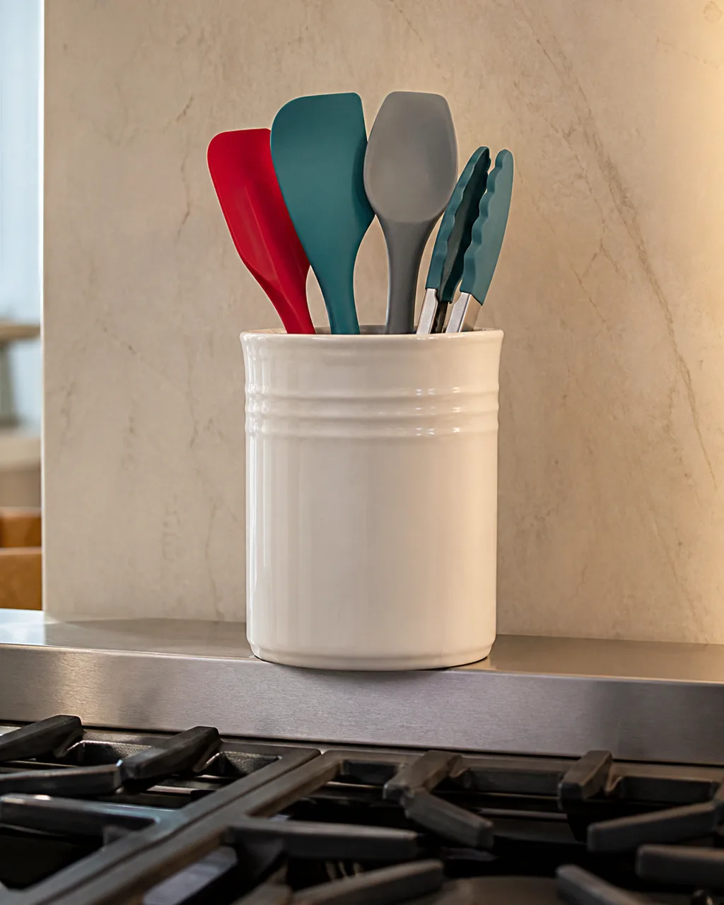 A white ceramic canister on the stainless steel apron behind gas stove burners holding colorful silicone spatulas and tongs upright with warm kitchen light