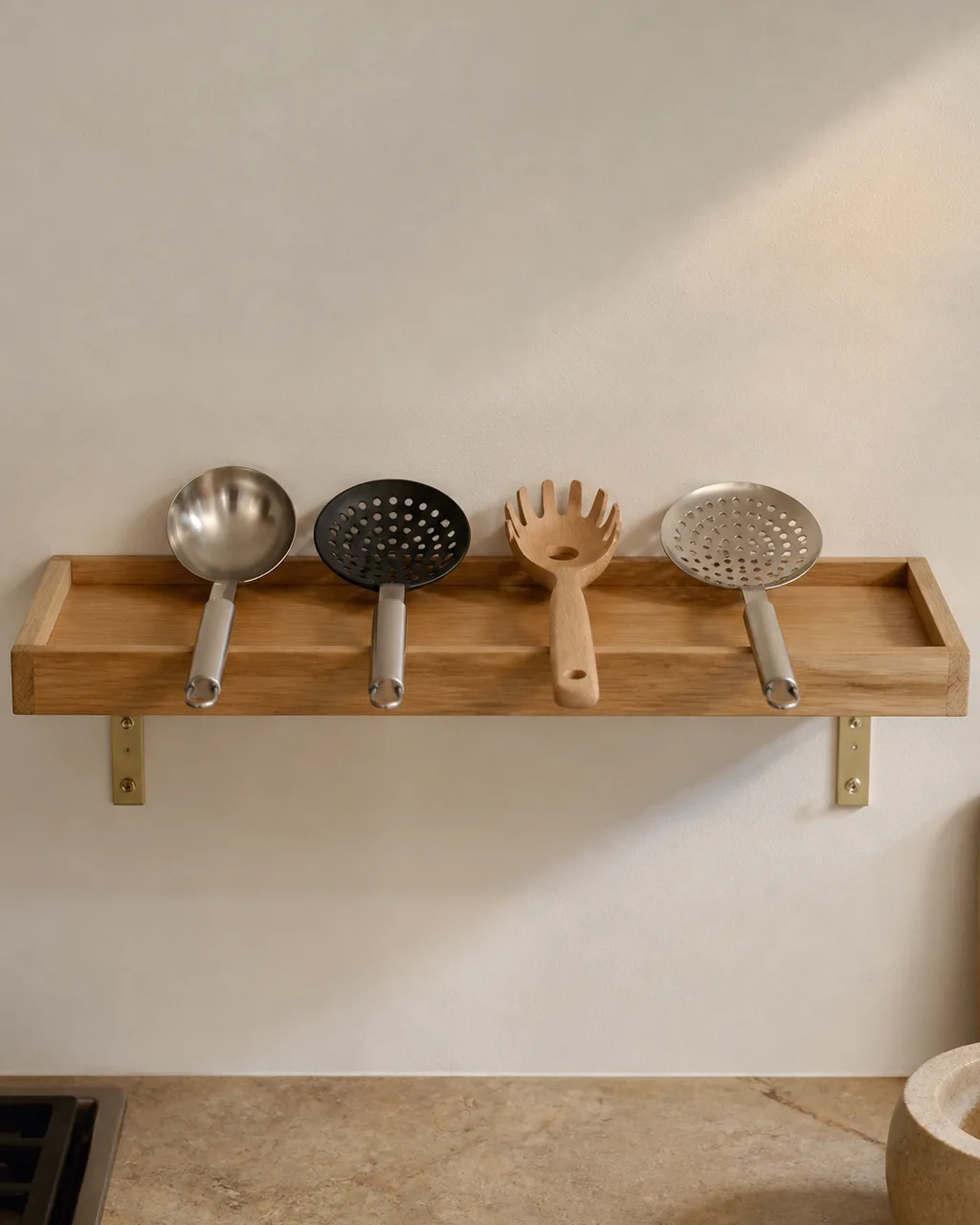 A narrow floating light oak tray on a white kitchen wall holding four long-handled utensils horizontally with visible brass mounting brackets