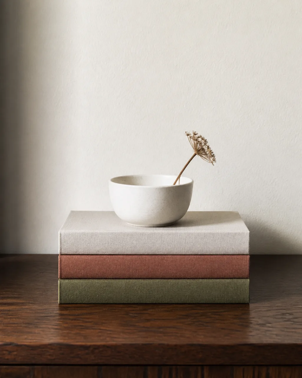 Three hardcover books stacked horizontally on a dark walnut console table with a small white ceramic bowl holding a dried flower stem on top