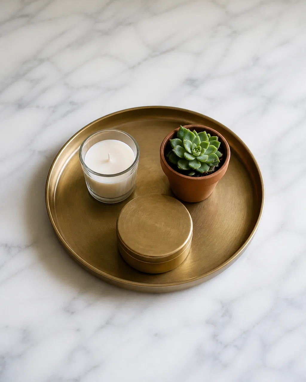 A round brass tray on a white marble console table holding a candle, a small succulent, and a decorative brass box arranged in a triangle